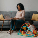 mulher negra, 30 anos, gravida, sentada no sofá cinza, à mesa própria para para laptop, trabalhando no laptop, enquanto uma criança de 3 anos brinca no chão da sala, sob um tapete de atividades.